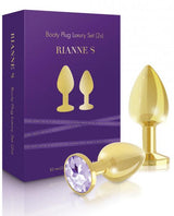 The Rianne S Booty Plug Set 2X Metal Gold, featuring a medical grade gold plug with a gem-like base, is shown next to a purple box labeled “Booty Plug Luxury Set (2x) RIANNE S,” which displays illustrations of two plugs.