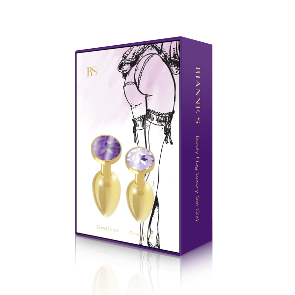 A box featuring the Rianne S Booty Plug Set 2X Metal Gold, a medical-grade gold butt plug set with purple gemstones, and packaging that displays an illustrated drawing of a lingerie-clad woman’s lower body.